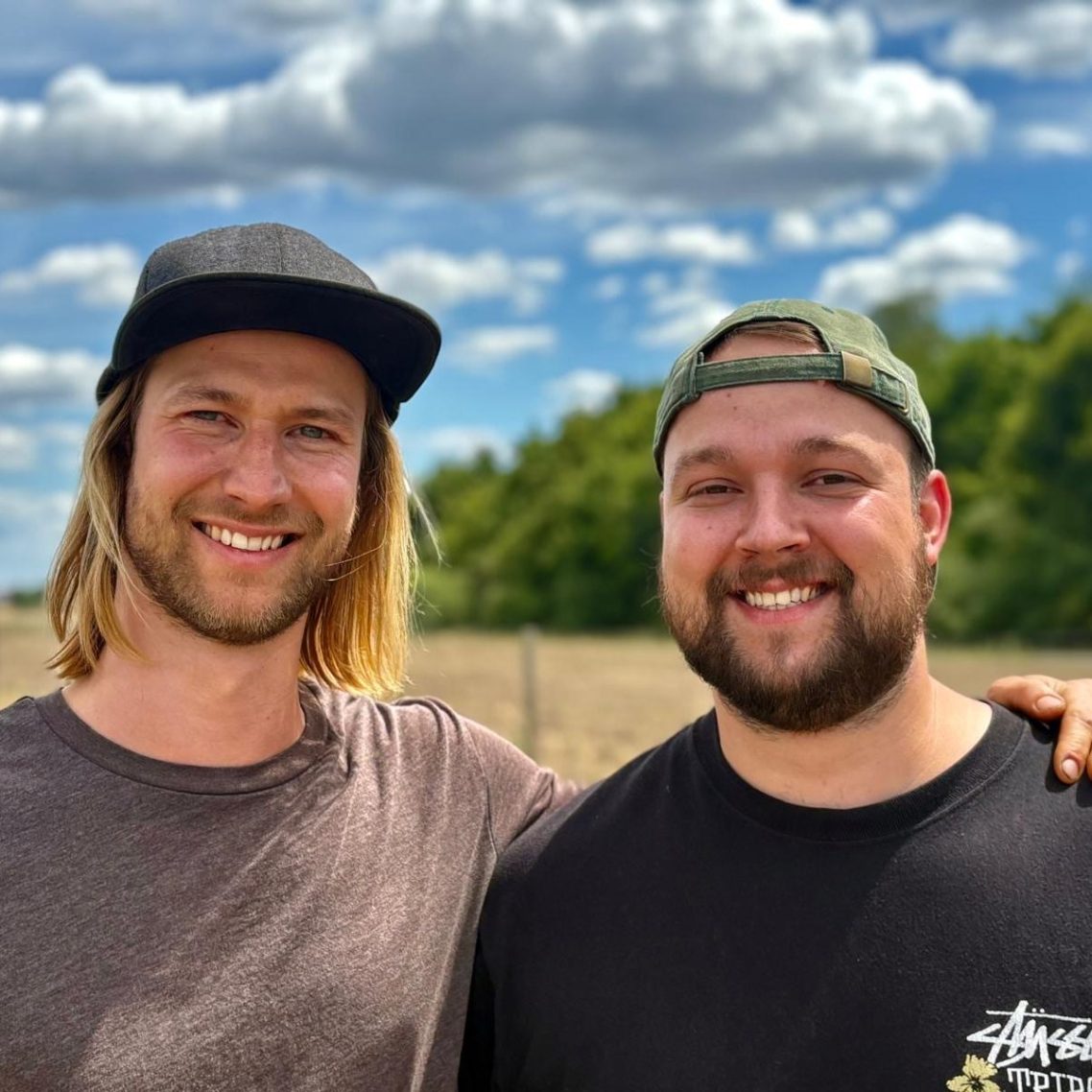 Yanik und Marek Rau Yanik und Marek Rau stehen lächelnd vor einer grünen Landschaft unter einem bewölkten Himmel.